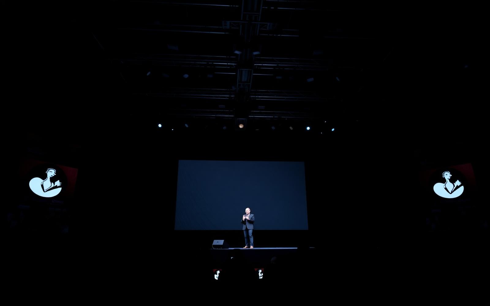 Keynote speaker on a dark conference stage with large LED screen